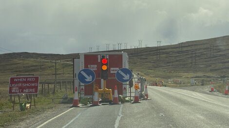 A temporary roadblock with a red traffic light and directional signs stops traffic on a rural road; a sign instructs drivers to wait when the red light shows.