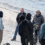 A group of people, some with cameras and equipment, stand and talk on a sandy beach near the water’s edge.