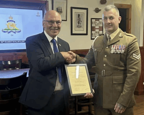Two men shake hands; one in a suit holds a certificate while the other, in military uniform with medals, faces the camera in a wood-paneled room.