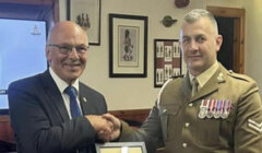 Two men shake hands indoors; one wears a suit and glasses, the other is in a decorated military uniform. Framed pictures are visible on the wall behind them.