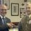 Two men shake hands indoors; one wears a suit and glasses, the other is in a decorated military uniform. Framed pictures are visible on the wall behind them.