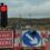 A red traffic light and road signs indicate construction ahead, with cones and vehicles blocking part of a rural road under cloudy skies.
