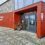 Entrance of Scalloway Museum with glass doors, a red exterior, a welcome sign, and potted plants beside the entrance.