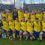 A men's soccer team in yellow and blue uniforms poses together in front of a goal, with coaches in dress shirts beside them. Two trophies are displayed on the grass in front.