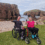 Two people in wheelchairs sit on grass by a rocky coastline, smiling and waving at the camera with cliffs and the sea in the background under a cloudy sky.