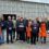 A group of people, some wearing orange aprons and name badges, pose together outdoors in front of a metal industrial building.