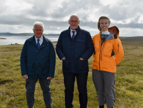 Three people stand outdoors on grassy terrain near a body of water under a cloudy sky, all wearing jackets and smiling at the camera.