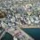 Aerial view of a coastal town with rows of gray buildings, a harbor with boats and cars, parking lots, and streets intersecting the residential and commercial areas.