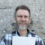 A man with a gray beard and mustache stands in front of a textured stone wall, holding a white piece of paper. He is wearing a plaid shirt and smiling.