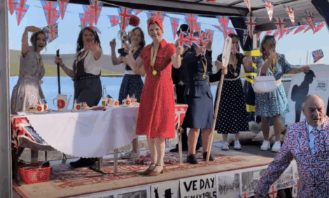 A group of people in vintage-style clothing stand on a decorated stage with Union Jack flags, waving and smiling during a VE Day celebration event.