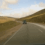 A car drives on a rural road through hilly terrain with wind turbines in the distance under a partly cloudy sky.