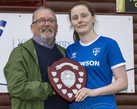 A man and a woman in a blue sports jersey hold a shield-shaped trophy together and smile at the camera.