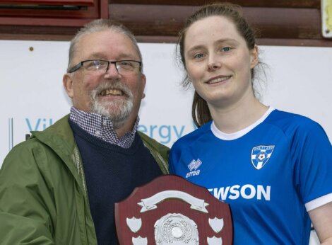 A man and a woman in a blue sports jersey hold a shield-shaped trophy together and smile at the camera.