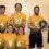 Six volleyball players in yellow-green uniforms pose indoors; one holds a shield-shaped trophy presented by a woman in black sportswear.