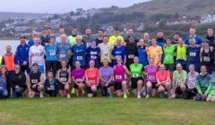A large group of people, including cyclists and runners with race bibs, pose together outdoors on grass near a body of water and houses in the background.
