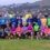 A large group of people, including cyclists and runners with race bibs, pose together outdoors on grass near a body of water and houses in the background.
