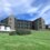 A modern three-story building with large windows and sloped roofs stands on a grassy area, with smaller buildings and bushes nearby under a partly cloudy sky.
