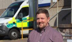 A man in a checked shirt stands outside a hospital building with an ambulance parked in the background.