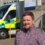 A man in a checked shirt stands outside a hospital building with an ambulance parked in the background.