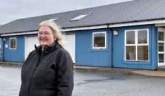 A person with long gray hair and glasses stands outside a blue building with white-framed windows and a gray roof, on a cloudy day.