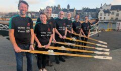 Seven people in matching black shirts stand in a row holding oars, with a boat and buildings visible in the background on a waterfront.