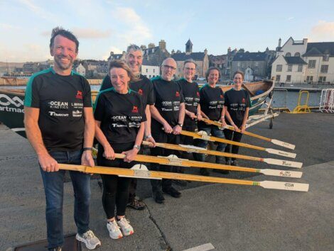 Seven people in matching black shirts stand in a row holding oars, with a boat and buildings visible in the background on a waterfront.