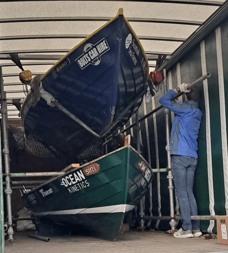 Two rowing boats are stacked inside a truck, with a person in a blue jacket using a pipe or tool near the boats.