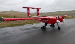 A red unmanned aerial vehicle with two propellers is parked on a wet paved surface near grassy fields under an overcast sky.