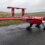 A red unmanned aerial vehicle with two propellers is parked on a wet paved surface near grassy fields under an overcast sky.