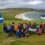 A group of people pose on a grassy hill by the coast, holding Scottish and Palestinian flags, with a beach and sea in the background under a cloudy sky.