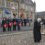 A group of people, some in military uniforms, stand in front of a stone building holding wreaths and flags during a remembrance ceremony. A person reads from a lectern while another stands in silence.