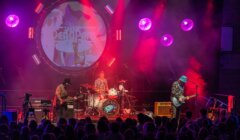Three musicians perform on stage with drums, guitar, and bass under colorful lighting, with "The Dead Pan" projected behind them. An audience watches from the foreground.