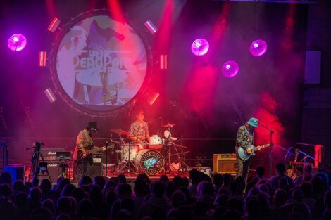 Three musicians perform on stage with drums, guitar, and bass under colorful lighting, with "The Dead Pan" projected behind them. An audience watches from the foreground.
