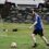 A man wearing a blue vest and black shorts is playing soccer on a grassy field, approaching a white and orange ball with cars and a stone wall in the background.