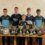 Seven boys in matching sports uniforms stand behind a table displaying numerous trophies and shields in a room with patterned wallpaper.