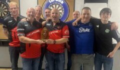 A group of eight men pose indoors near dartboards; two of them at the front are holding a trophy and wearing matching red and black shirts.