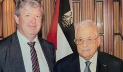 Two men in suits stand side by side in front of a flag with Egyptian colors inside a formal room.