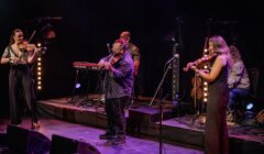 Four musicians perform on stage with violins, a keyboard, and microphones under stage lights, in front of a dark background.
