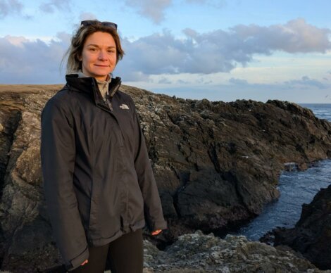 A woman wearing a dark jacket stands on rocky terrain near the edge of the sea under a mostly cloudy sky.