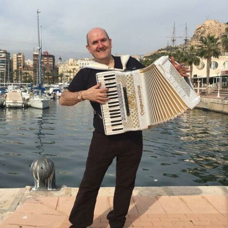 A man stands by a marina playing a large accordion, with boats, buildings, and palm trees visible in the background.