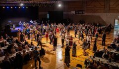 A large group of people dance and socialize in a gymnasium with a stage and live band, while others sit at tables along the sides.
