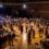 A large group of people dance and socialize in a gymnasium with a stage and live band, while others sit at tables along the sides.