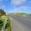 A wide road with multiple lanes runs through a rural area, bordered by grass and some construction materials, under a blue sky with scattered clouds.