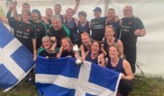 A group of people celebrate indoors holding trophies and Scottish flags, smiling and wearing athletic clothing.