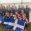 A group of people celebrate indoors holding trophies and Scottish flags, smiling and wearing athletic clothing.