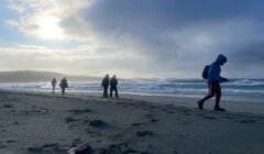 Several people walk along a cloudy, windswept beach near the ocean, with footprints visible in the sand and waves in the background.
