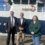 Three adults stand side by side on a dock in front of a large white and blue boat. The two men wear suits, and the woman wears a green patterned top.