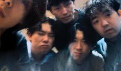 Five young people pose closely together for a group selfie indoors, all looking toward the camera with neutral or slightly serious expressions.