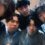 Five young people pose closely together for a group selfie indoors, all looking toward the camera with neutral or slightly serious expressions.