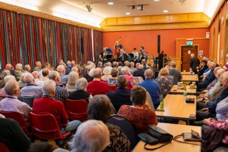 A seated audience watches a small music ensemble perform on stage in a brightly lit room with colorful curtains.